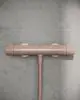 Brausethermostat in Rosé von Ideal Standard, mit zwei verstellbaren Griffen, montiert an einer grauen Wand. Der Thermostat hat einen geraden Wasserzu- und -abgang.